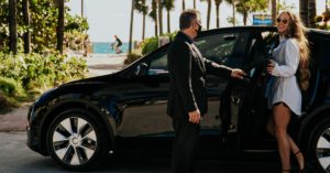 A woman being picked up by a Presidential Limo vehicle in Miami. The driver is opening the back door for her. 